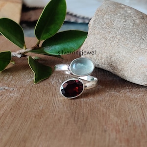 May include: A silver ring with a light blue oval gemstone and a dark red oval gemstone. The ring is on a wooden surface with green leaves and a rock in the background.