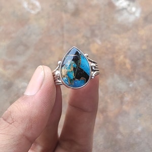May include: Silver ring with a teardrop-shaped stone. The stone has a mix of blue, black, and gold colours. The ring has a detailed band with small decorative elements. The ring is held by a hand.