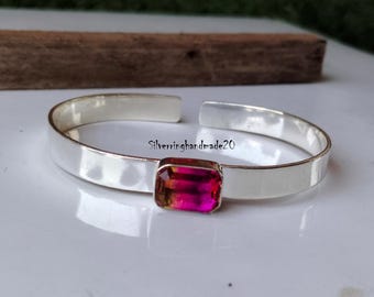 May include: A silver bangle bracelet with a pink and yellow rectangular gemstone set in the center. The bracelet is open and has a smooth, polished finish.