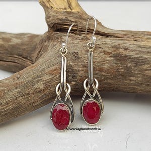 May include: A pair of silver dangle earrings with red oval gemstones. The earrings feature a delicate knot design.