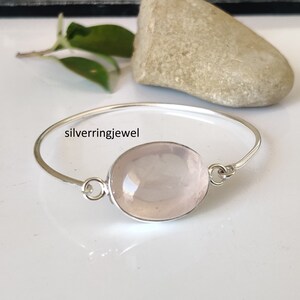 Könnte beinhalten: Ein silbernes Armkettchen mit einem großen ovalen Rosenquarz-Edelstein. Das Armband ist offen und hat ein schlichtes Design.