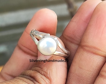 May include: A silver ring with a large, round, white pearl. The ring has a split band design, with the pearl held in place by a bezel setting. The ring is held in a hand, with the text "Silverringhandmade20" visible.