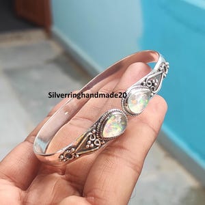 Puede incluir: Un brazalete de plata con dos ópalos iridiscentes en forma de pera engastados en metal ornamentado. El brazalete tiene un acabado pulido y detalles decorativos intrincados alrededor de las piedras. El texto Silverringhandmade20 es visible.