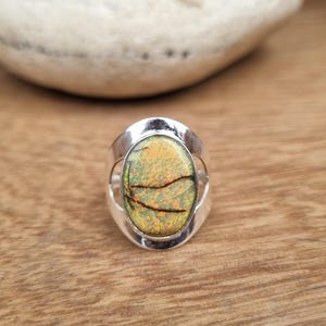 May include: A silver ring with an oval-shaped gemstone. The gemstone is a light green and yellow color with brown veins running through it.