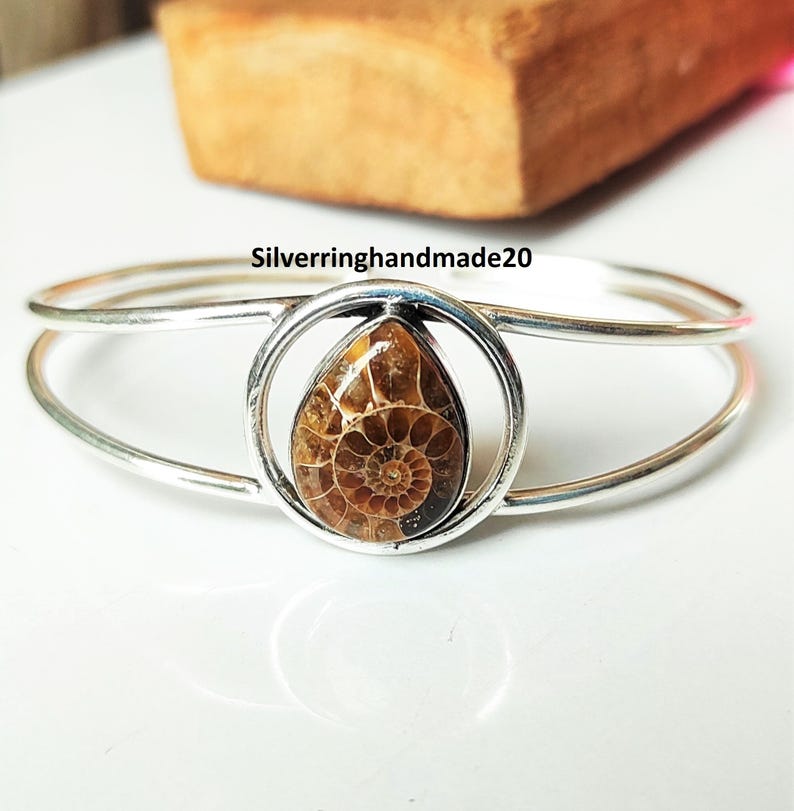 May include: A silver-toned bracelet featuring a teardrop-shaped, fossilized ammonite stone. The stone is encased in a circular setting, with the text "Silverringhandmade20" visible. The bracelet has a double-band design.
