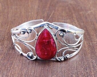 May include: A silver bangle bracelet featuring a large, teardrop-shaped red gemstone set in the centre. The bracelet has a delicate, ornate design with swirling patterns.