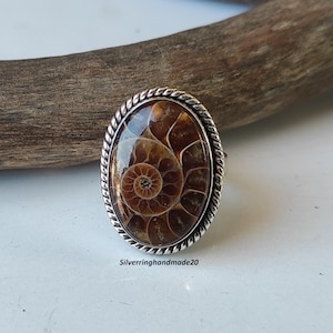 May include: A silver ring with an oval-shaped ammonite fossil gemstone. The fossil is brown and has a spiral pattern. The ring has a rope-like design around the edge.
