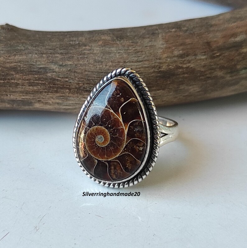 May include: A silver ring with a brown ammonite gemstone set in a teardrop-shaped bezel. The ring has a rope-like design around the bezel and a silver band.