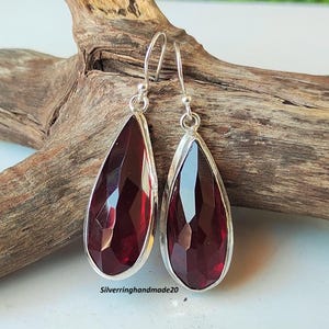 May include: A pair of silver dangle earrings with teardrop-shaped red gemstone accents. The earrings are on a wooden surface.