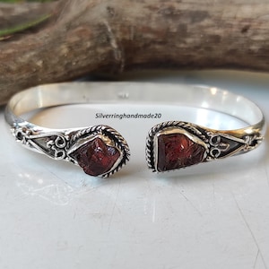 Puede incluir: Pulsera de plata con dos piedras de granate rojo engastadas en plata. La pulsera tiene un diseño delicado con un borde de cuerda y detalles florales.