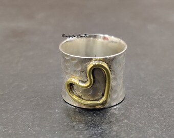 Può includere: Un anello d'argento con una texture martellata e un design a forma di cuore dorato. L'anello è largo e ha una forma arrotondata.
