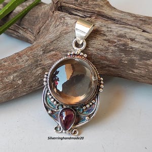 Può includere: Un ciondolo d'argento con una grande pietra di quarzo trasparente e una pietra di granato più piccola. Il ciondolo ha un design intricato con perline d'argento e una finitura smaltata blu e verde.