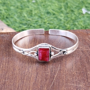 Può includere: Un braccialetto rigido in argento con una pietra preziosa rossa incastonata in una cornice d'argento. Il braccialetto ha un design delicato e decorato.