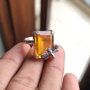 Könnte beinhalten: Ein silberner Ring mit einem großen, rechteckigen, orangefarbenen Edelstein und einem kleinen Bienenmotiv.