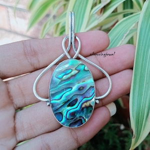 Könnte beinhalten: Ein silberner Anhänger mit einem ovalen Einlegeteil aus Abalone-Muschel. Der Anhänger hat ein geknotetes Design und eine silberne Öse.