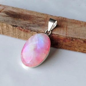 Puede incluir: Un colgante de plata con una gran piedra de luna rosa ovalada. La piedra de luna tiene un brillo iridiscente arcoíris.