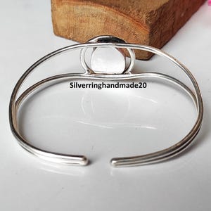 May include: A silver-toned cuff bracelet with a central, circular, light-colored stone. The bracelet has a simple, open design with the text "Silverringhandmade20" visible on the surface. The bracelet is displayed on a light-colored surface.