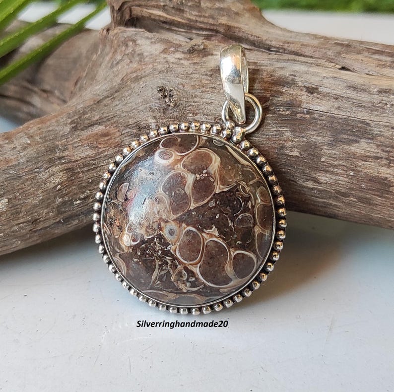 May include: A round pendant with a brown and white agate stone set in a silver bezel. The pendant has a beaded edge and a silver bail.