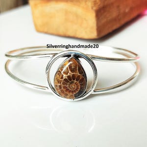 May include: Silver-toned bracelet featuring a teardrop-shaped, fossilized ammonite. The ammonite is encased in a circular setting, with the bracelet's band curving around the wrist. The text "Silverringhandmade20" is visible.