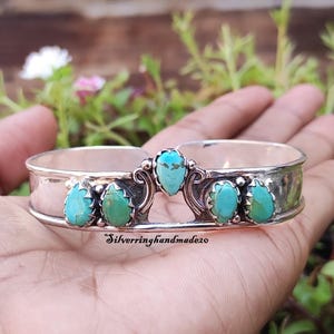May include: A silver cuff bracelet featuring five teardrop-shaped turquoise stones set in scalloped bezels. The centre stone is elevated above the others, flanked by decorative silver scrollwork. The text Silverringhandmade20 is visible.