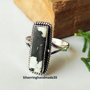 May include: A silver ring with a rectangular black and white stone. The stone is set in a decorative frame with a twisted rope design. The ring has a split shank and is displayed on a white surface.