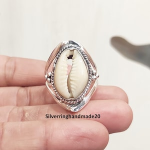 May include: A silver ring with a cream-coloured cowrie shell centrepiece. The shell is set within a decorative silver frame with a twisted rope design. The ring has a unique, handcrafted appearance.