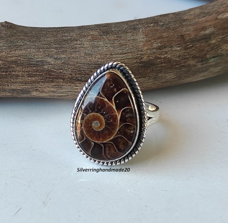 May include: A silver ring with a teardrop-shaped ammonite gemstone. The ring has a braided silver band and a detailed silver setting around the gemstone.