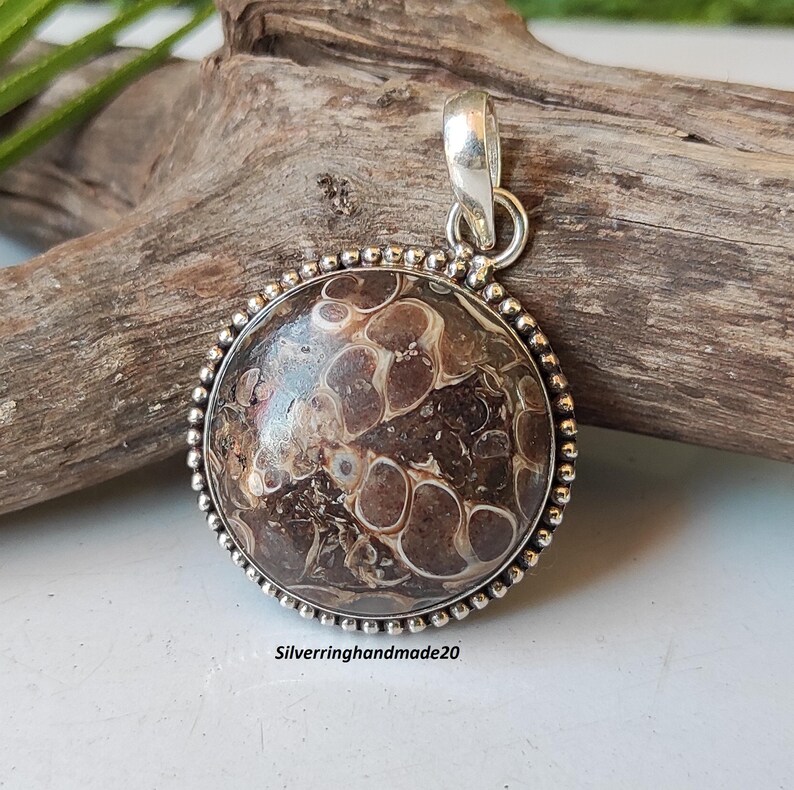 May include: A round pendant with a brown and white agate stone set in a silver bezel. The pendant has a beaded edge and a silver bail.