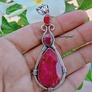Puede incluir: Un colgante de plata con una gran piedra preciosa roja en forma de lágrima rodeada de piedras preciosas rojas más pequeñas. El colgante está engastado en un diseño de alambre de plata.