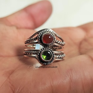 May include: Two silver rings with a gemstone design. One ring features a red gemstone, and the other has a green gemstone. The rings have a twisted rope design around the gemstones. The text "Silverringhandmade20" is visible.