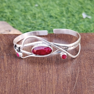 Könnte beinhalten: Ein silbernes Armreif-Armband mit einem roten Edelstein-Design. Das Armband verfügt über einen großen ovalen roten Edelstein in der Mitte und zwei kleinere runde rote Edelsteine ​​auf beiden Seiten. Das Armband besteht aus einem filigranen Silberdraht, der verdreht und ineinander verschlungen ist, um ein einzigartiges und kompliziertes Design zu schaffen.