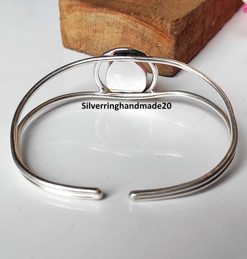 May include: A silver-toned cuff bracelet with a central, circular, light-colored stone. The bracelet has a simple, open design with the text "Silverringhandmade20" visible on the surface. The bracelet is displayed on a light-colored surface.
