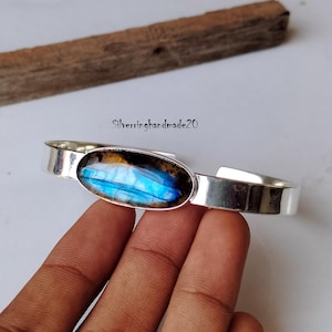 May include: A silver cuff bracelet featuring a large oval-shaped labradorite gemstone set in the centre. The gemstone displays a blue and green iridescent sheen.