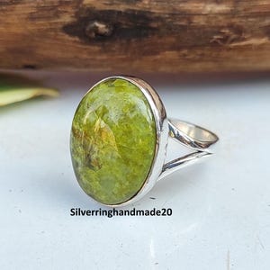 Peut inclure: Bague en argent avec une grosse pierre ovale vert clair. La pierre a une surface texturée avec des variations de couleur. Le corps de la bague se divise en deux brins. Le texte "Silverringhandmade20" est visible. Bijou.