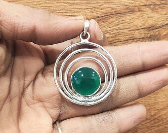 May include: A silver pendant with a circular design. The pendant features three concentric rings surrounding a vibrant, round, green gemstone. The gemstone is the focal point, set in the center of the design. The pendant is held in a hand.