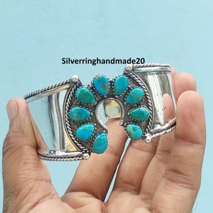 May include: A silver cuff bracelet featuring a central horseshoe-shaped design adorned with eight teardrop-shaped turquoise stones. The metal band has a polished finish with twisted rope detailing along the edges. The text Silverringhandmade20 is visible.