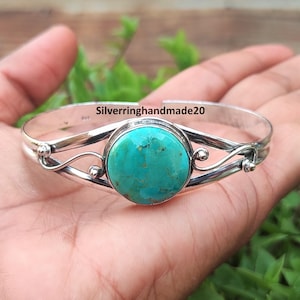 May include: A silver cuff bracelet featuring a round, polished turquoise stone at the centre. The metal band displays a swirling, open-work design with small spherical accents on each side. The text Silverringhandmade20 is visible on the band.