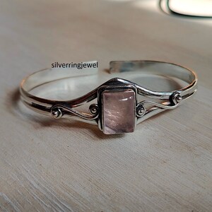 Könnte beinhalten: Ein silbernes Armreif-Armband mit einem zarten, wirbelnden Design. Das Armband hat einen rechteckigen rosa Edelstein in der Mitte.
