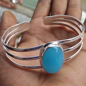 May include: A silver bangle bracelet with a light blue oval gemstone. The bracelet has three bands and is open on one side.