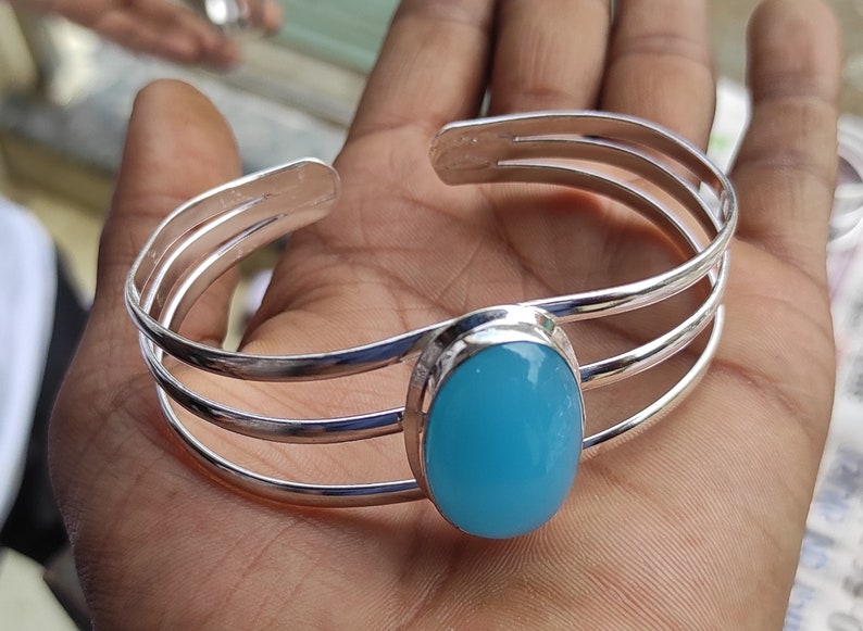 May include: A silver bangle bracelet with a light blue oval gemstone. The bracelet has three bands and is open on one side.