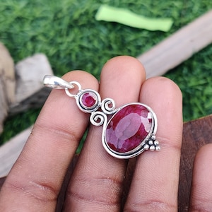 以下が含まれることがあります： 大きな楕円形のルビーと、小さな丸いルビー2つがついたシルバーのペンダント。ペンダントは繊細で装飾的なデザインです。