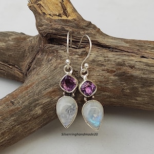 May include: A pair of silver earrings with a teardrop-shaped moonstone and a square amethyst gemstone. The earrings have a simple design and are perfect for everyday wear.