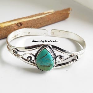 May include: A silver cuff bracelet featuring a central pear-shaped turquoise stone with dark veining. The metal band displays intricate swirling wirework patterns on both sides of the stone. The text "Silverringhandmade20" is visible on the band.