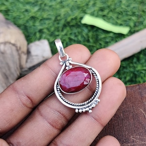 Puede incluir: Un colgante de plata con una piedra preciosa roja engastada en un marco circular. El colgante tiene un aro para sujetarlo a una cadena.