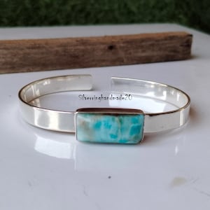 Könnte beinhalten: Ein silbernes Armreif-Armband mit einem rechteckigen blauen und weißen Edelstein. Das Armband ist offen und hat ein schlichtes Design.