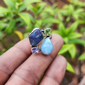 May include: A silver ring featuring a blue, green, and light blue gemstone setting. The ring is set with a large blue gemstone, a smaller green gemstone, and a teardrop-shaped light blue gemstone.