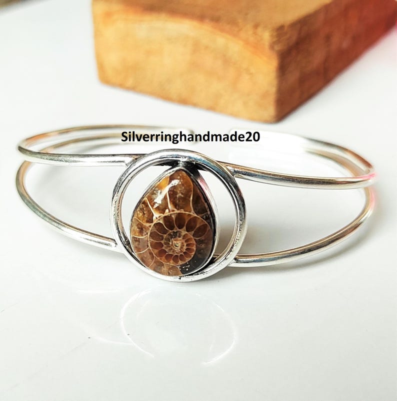 May include: A silver-tone bracelet featuring a teardrop-shaped, fossilized ammonite stone. The stone is encased in a circular setting, with the text "Silverringhandmade20" visible on the bracelet. The bracelet has a double-band design.