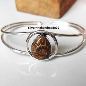 May include: Silver cuff bracelet featuring a teardrop-shaped fossil set in a circular frame. The fossil displays a detailed spiral pattern in shades of brown. The bracelet has a double-band design and the text "Silverringhandmade20".