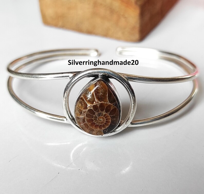 May include: Silver cuff bracelet featuring a teardrop-shaped fossil set in a circular frame. The fossil displays a detailed spiral pattern in shades of brown. The bracelet has a double-band design and the text "Silverringhandmade20".