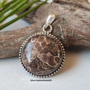 May include: A silver pendant with a round, brown and white agate stone. The stone has a marbled pattern. The pendant is on a wooden surface.